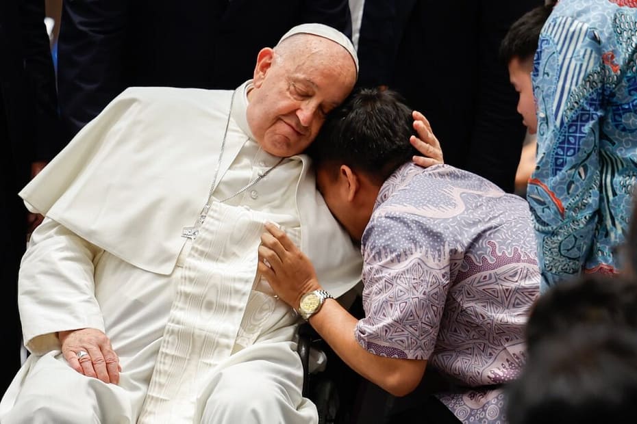 A man leans emotionally on the shoulder of a seated Pope Francis in white robes and a skullcap, surrounded by other people.